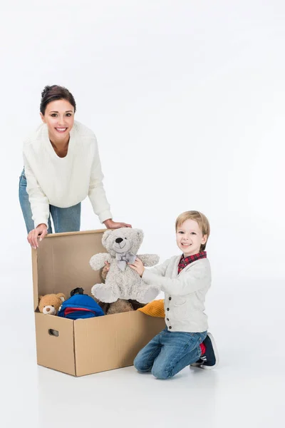 Happy mother with son holding teddy bear and sitting near cardboard box with toys isolated on white — Stock Photo