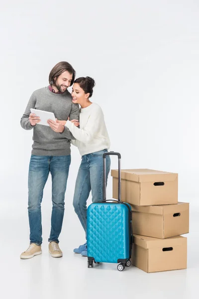 Heureux couple en utilisant tablette numérique debout près de valise bleue et boîtes en carton isolés sur blanc — Photo de stock