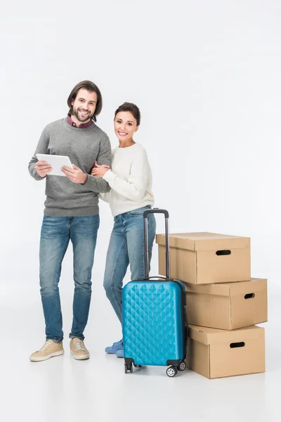 Улыбающаяся пара с помощью цифровой планшет стоя рядом с чемоданом и картонные коробки изолированы на белом — стоковое фото