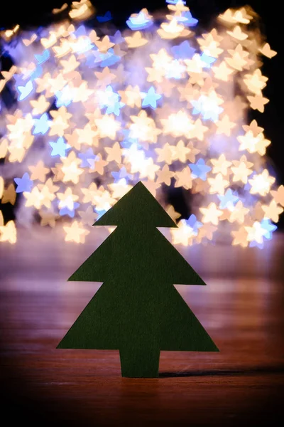 Close up view of green paper christmas tree on wooden tabletop and bokeh lights in shape of stars background — Stock Photo