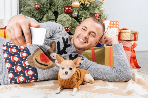 Lächelnder Mann macht Selfie mit Chihuahua-Hund mit Weihnachtsbaum zu Hause — Stockfoto