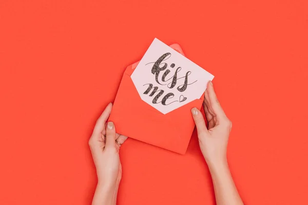 Cropped shot of person holding red envelope with card and kiss me inscription isolated on red — Stock Photo