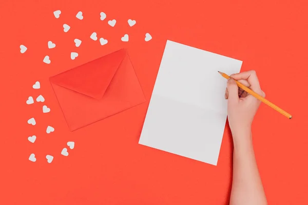 Cropped shot of person writing in white card, red envelope and small hearts isolated on red — Stock Photo
