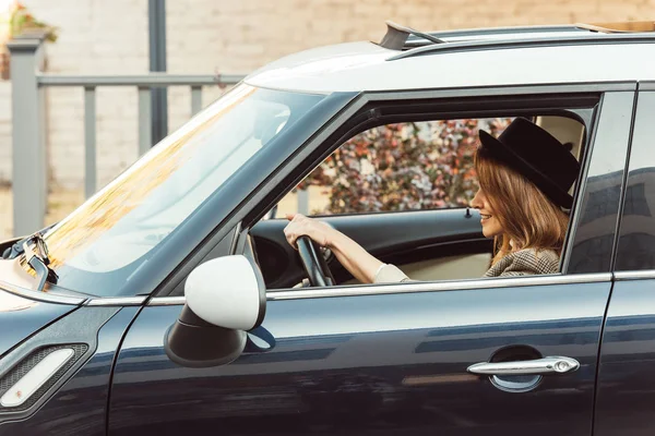 Seitenansicht einer lächelnden stilvollen Frau in karierter Jacke und schwarzem Hut, die Auto fährt — Stockfoto