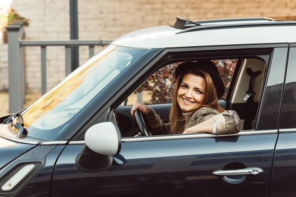 Donna allegra in giacca a scacchi e cappello nero seduto in auto in strada urbana — Foto stock