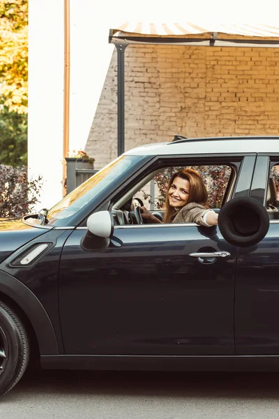 Smiling woman leaning out hand with black hat while sitting in car at urban street — Stock Photo