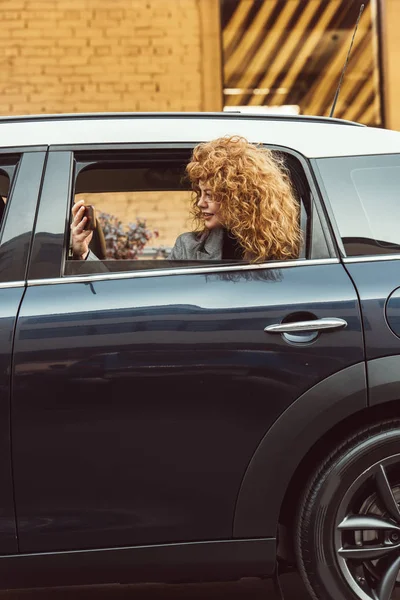 Vue latérale de la rousse bouclée femme penchée hors de la voiture et de prendre selfie sur smartphone — Photo de stock