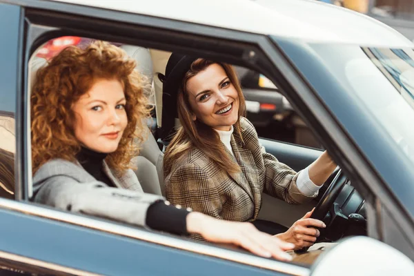 Selective focus of smiling stylish woman in black hat driving car while her ginger female friend sitting near — Stock Photo