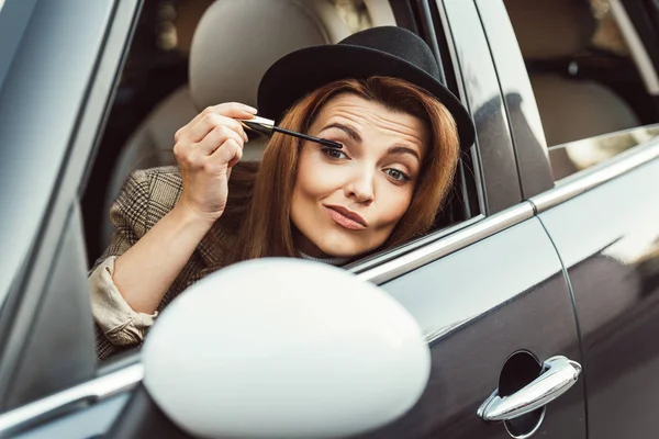 Красивая женщина в черной шляпе, смотрящая на боковое зеркало, используя чернила для ресниц в машине — стоковое фото