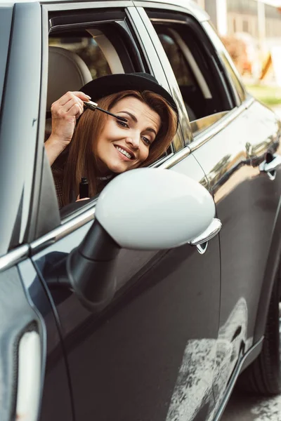 Щаслива доросла жінка в чорному капелюсі дивиться на дзеркало крила під час макіяжу в машині — стокове фото