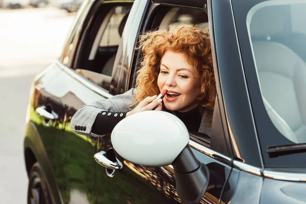 Mujer jengibre feliz mirando el espejo del ala mientras se utiliza lápiz labial rojo en el coche - foto de stock