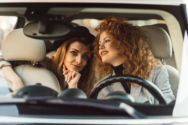 Souriant roux femme parler à un ami féminin alors qu'elle est assise sur le siège arrière de la voiture — Photo de stock