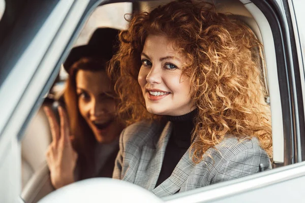 Portrait de gingembre gai femme regardant la caméra tandis que son amie montrant signe de victoire dans la voiture — Photo de stock