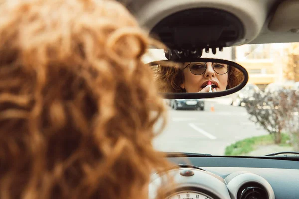 Vista parcial de mujer pelirroja en anteojos mirando el espejo trasero y usando lápiz labial rojo en el coche - foto de stock