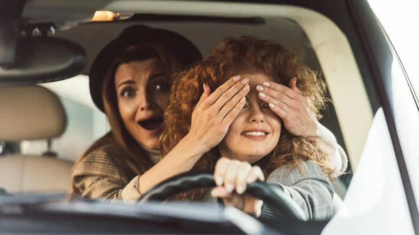Selective focus of shocked woman covering eyes to female friend while she driving car — Stock Photo