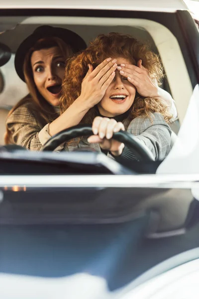 Shocked woman covering eyes to female friend while she driving car — Stock Photo