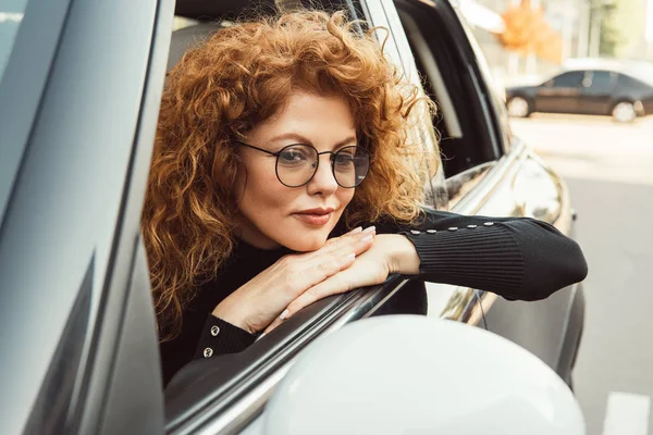 Селективный фокус рыжеволосой рыжей женщины в очках, смотрящей на зеркало крыла в машине — стоковое фото