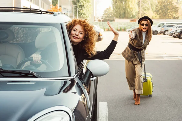 Redhead curly woman driving away and waving by hand to stylish female tourist with wheeled bag at city street — Stock Photo