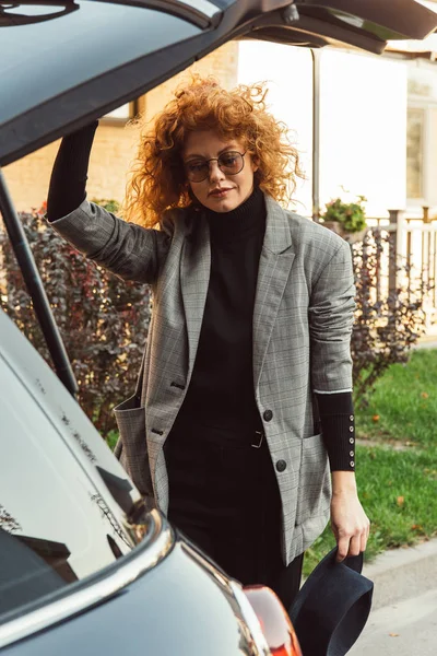 Mujer pelirroja rizada con estilo en chaqueta gris y gafas cerrando el maletero del coche en la calle de la ciudad - foto de stock