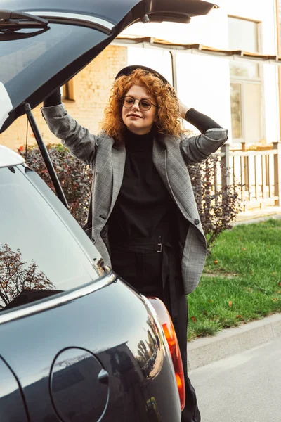 Élégante rousse bouclée femme en veste grise fermeture coffre de voiture à la rue de la ville — Photo de stock
