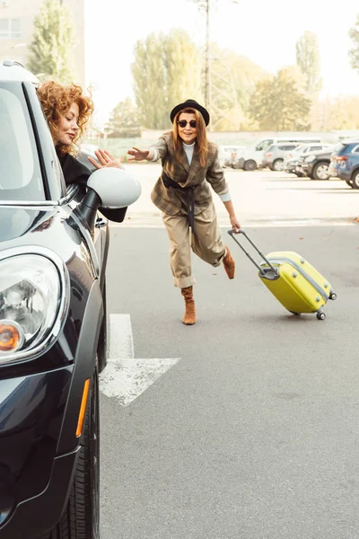 Femme heureuse conduisant loin et agitant à la main à touriste féminine élégante avec sac à roulettes à la rue de la ville — Photo de stock