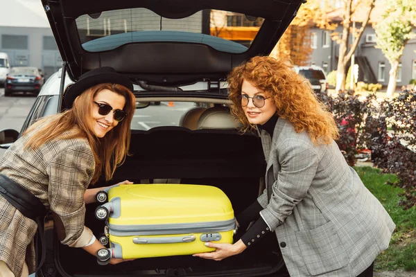 Belles touristes féminines élégantes en lunettes de soleil et vestes mettre sac à roulettes dans le coffre de la voiture à la rue de la ville — Photo de stock