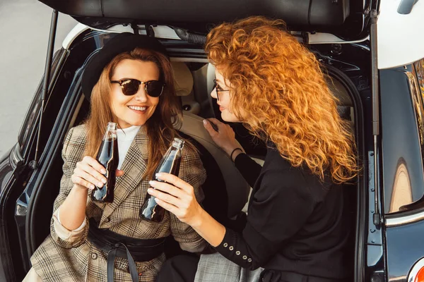 Femmes adultes souriantes en vestes cliquetis par des bouteilles de soda dans le coffre de la voiture dans la rue urbaine — Photo de stock
