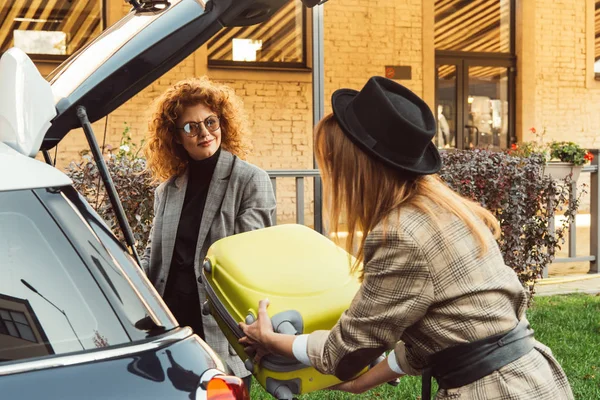 Rear view of female tourist putting wheeled bag into car trunk while ginger woman standing near at city street — Stock Photo