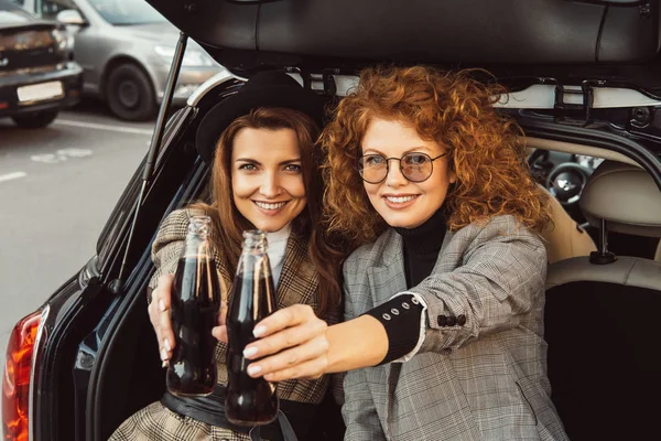 Feliz amigos do sexo feminino clinking por garrafas de refrigerante no porta-malas do carro na rua urbana — Fotografia de Stock