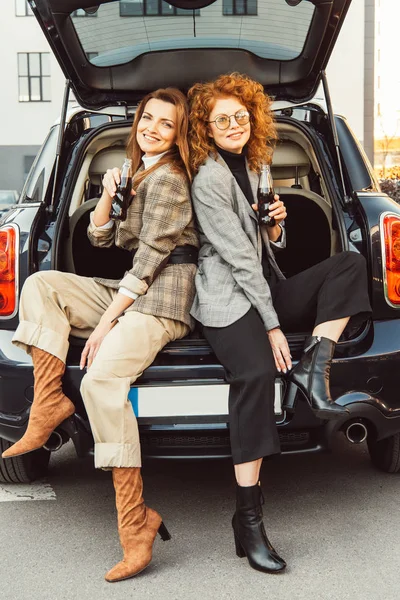 Cheerful female friends in jackets posing with soda bottles and sitting back to back in car trunk at urban street — Stock Photo