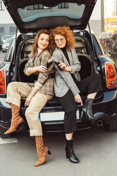 Stylish female friends in jackets clinking by soda bottles in car trunk at urban street — Stock Photo