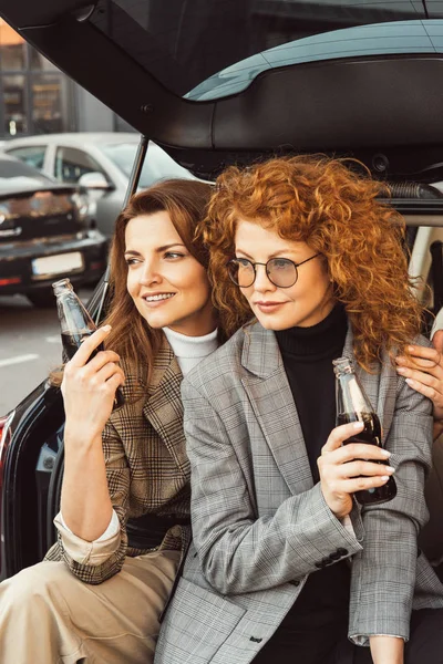 Mulheres elegantes em jaquetas olhando para longe e posando com garrafas de refrigerante no porta-malas do carro na rua urbana — Fotografia de Stock