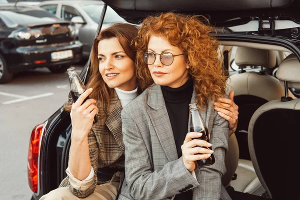 Selective focus of fashionable female models in jackets posing with soda bottles in car trunk at urban street — Stock Photo