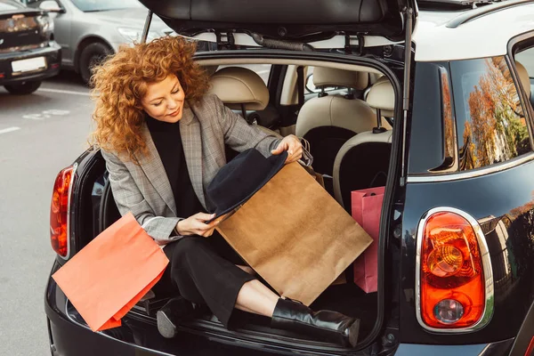 Elegante pelirroja rizado mujer con sombrero negro y bolsas de compras sentado en el maletero del coche - foto de stock