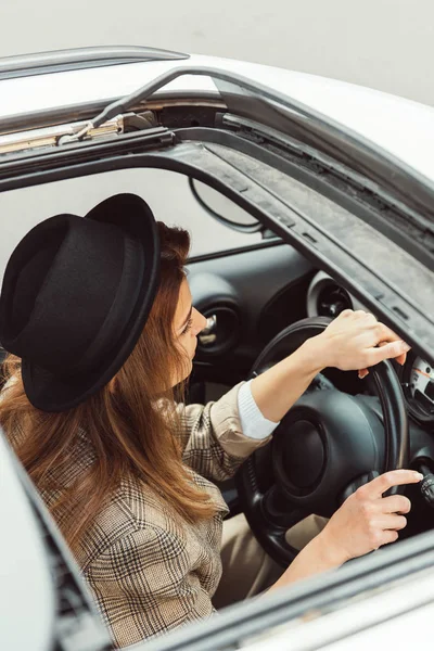 High angle view of fashionable woman in black hat and checkered jacket sitting at steering wheel in car — Stock Photo