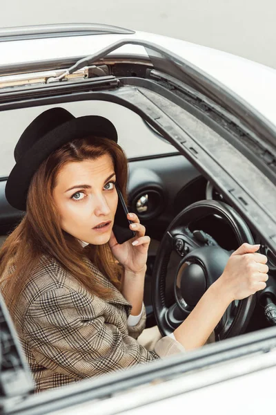 Retrato de mujer con estilo en sombrero negro hablando en el teléfono inteligente mientras está sentado en el volante en el coche - foto de stock