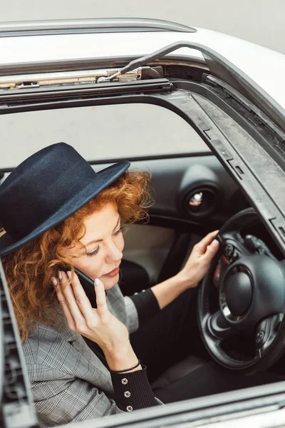 Vista de ángulo alto de la mujer pelirroja en sombrero negro hablando en el teléfono inteligente mientras está sentado en el volante en el coche - foto de stock