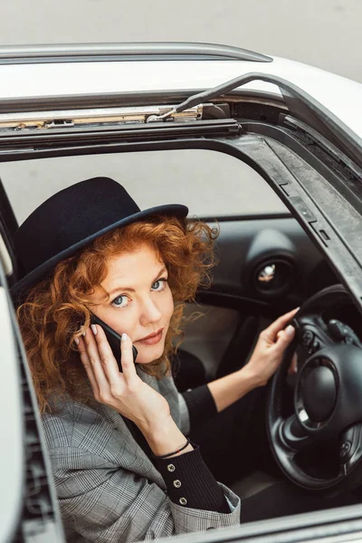 Vista de ángulo alto de la atractiva mujer de jengibre en sombrero negro hablando en el teléfono inteligente mientras está sentado en el volante en el coche - foto de stock