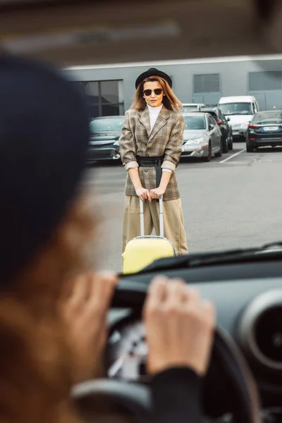 Imagem cortada de mulher sentada no volante enquanto turista elegante feminino de pé com saco de rodas na rua da cidade — Fotografia de Stock