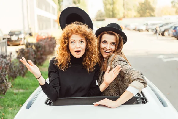 Happy woman taking off black hat from her surprised female friend while they leaning out from car hatchway at city street — Stock Photo