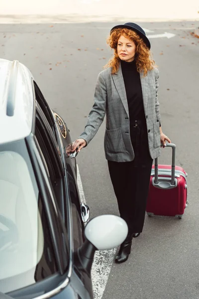 Vista ad alto angolo di rossa elegante viaggiatore femminile che trasporta borsa a ruote e porta di apertura di auto in strada urbana — Foto stock