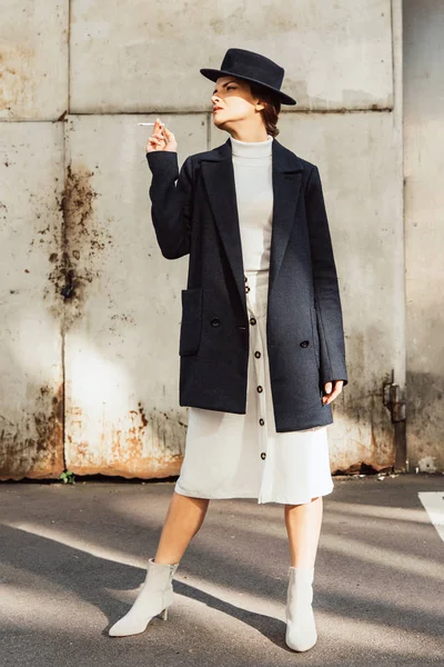 Modelo feminino elegante em casaco e chapéu preto posando com cigarro na mão na rua urbana — Fotografia de Stock