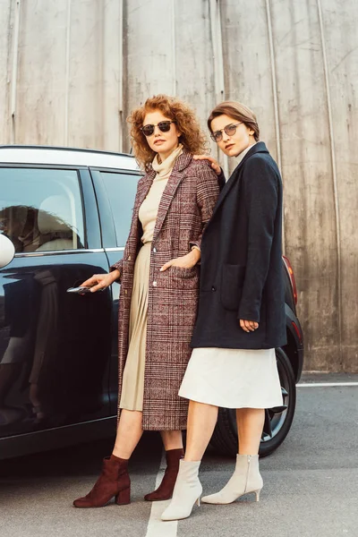 Sonrientes mujeres de moda en abrigos y gafas de sol posando cerca de coche en la calle de la ciudad - foto de stock