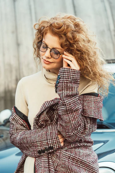 Selective focus of stylish ginger woman adjusting eyeglasses near car at urban street — Stock Photo