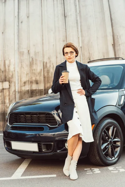 Женщина в пальто и очках, стоящая с одноразовой чашкой кофе возле машины на городской улице — стоковое фото
