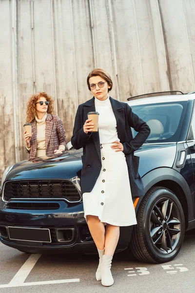 Modèles féminins à la mode en manteaux posant avec des tasses à café jetables près de la voiture à la rue urbaine — Photo de stock