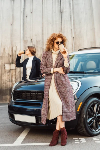 Modelos femeninas adultas en abrigos bebiendo café cerca del coche en la calle urbana - foto de stock