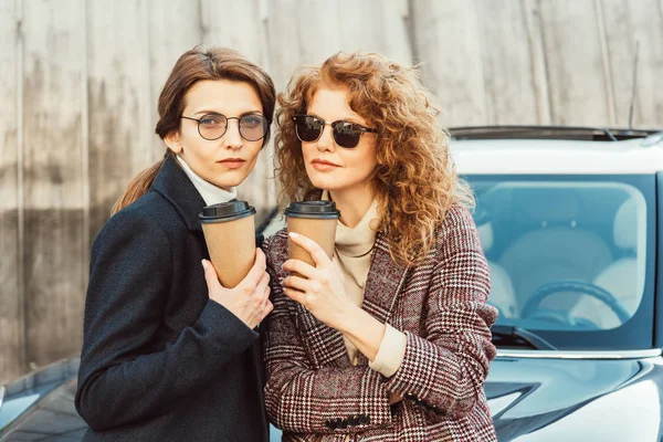 Selective focus of fashionable female friends in sunglasses and coats drinking coffee near car at urban street — Stock Photo