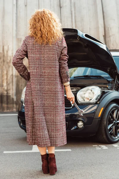 Rückansicht der stilvollen Ingwerfrau im Mantel mit Schraubenschlüssel in der Nähe des Autos an der Stadtstraße — Stockfoto