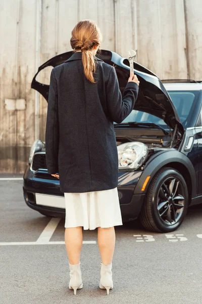 Задний вид женщины в пальто стоящей с гаечным ключом рядом с автомобилем с открытым капотом на улице города — стоковое фото